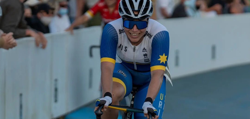 Australian cyclist wearing a blue and white uniform, racing on a track with a crowd in the background.